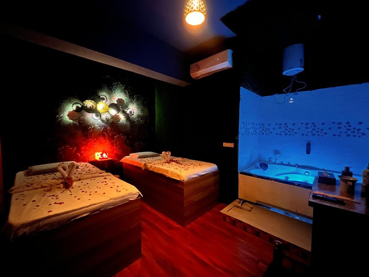 Couple spa suite — two beds with rose petals, jacuzzi glowing blue and red candlelight ambiance